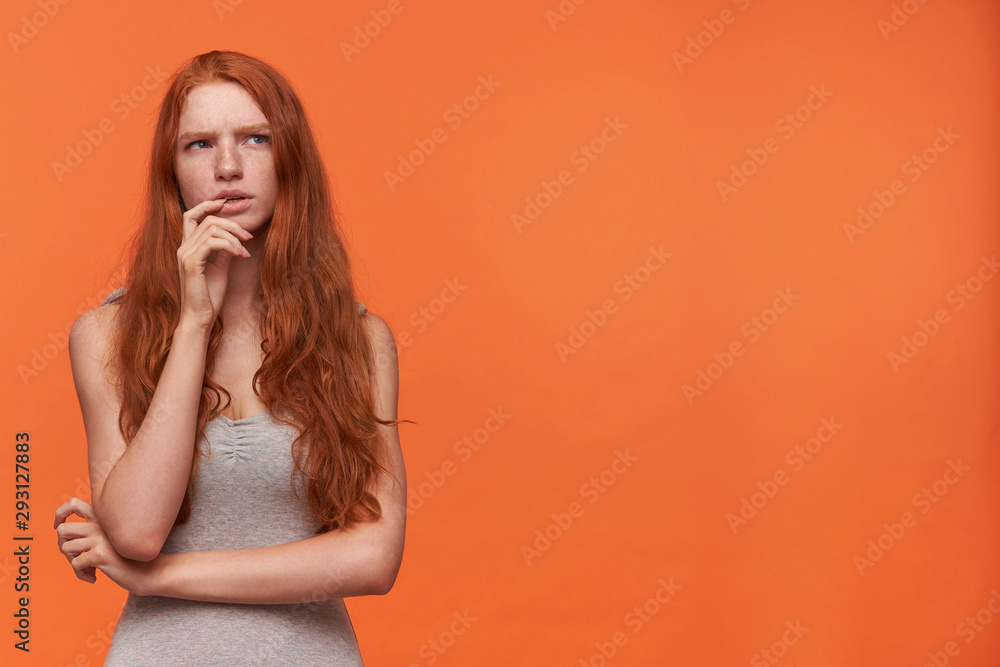 Studio shot of young attractive red haired female in casual clothes standing over orange background, holding chin with hand and looking aside pensively, trying to gather with thoughts Studio shot of young attractive red haired female in casual clothes standing over orange background, holding chin with hand and looking aside pensively, trying to gather with thoughts