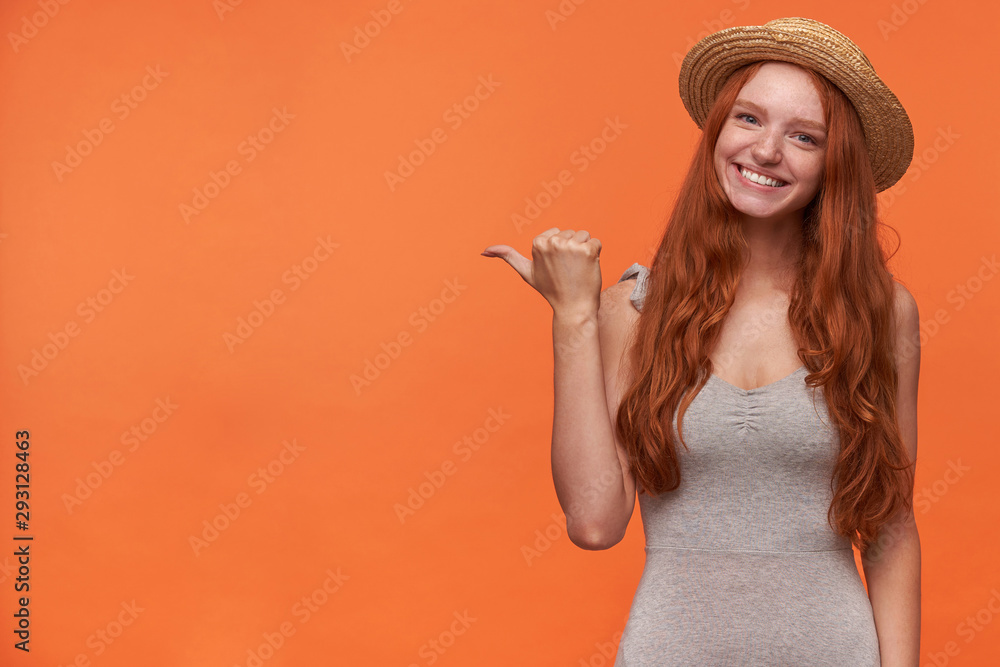 Portrait of positive young foxy lond haired female looking to camera with charming smile, standing over orange background with raised thumb showing aside, wearing casual clothes Portrait of positive young foxy lond haired female looking to camera with charming smile, standing over orange background with raised thumb showing aside, wearing casual clothes