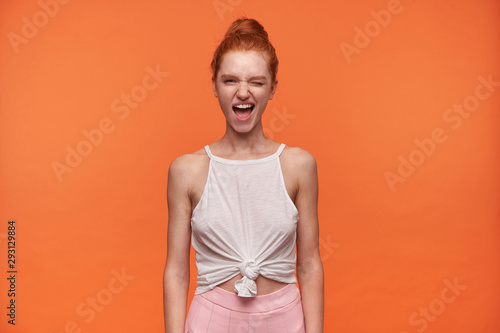 Indoor shot of coquettish young woman wearing her red hair in knot, posing over orange background in casual clothes with hands down, giving a wink to camera with wide mouth opened