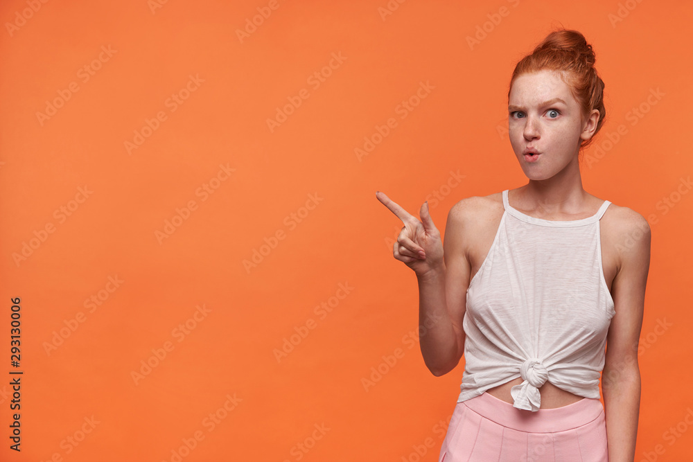 Obraz premium Horizontal shot of pretty young redhead woman with bun hairstyle wearing casual clothes, looking to camera and moving eyebrows, pointing away with forefinger, isolated over orange background