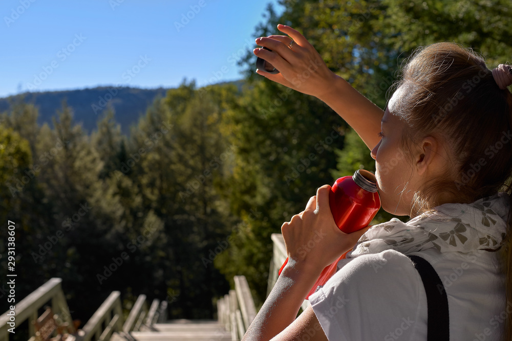 Naklejka premium Attractive young blonde in a white T-shirt drinking water from a bottle, standing in the mountains on a sunny summer day. Retouching, bright colors