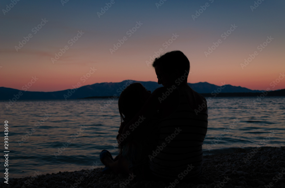 Mother holding her baby girl in her lap, sitting on a beach after sunset.