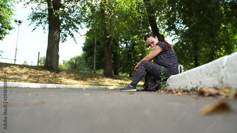 A pretty young woman in a sports suit with long hair sits on the curb and looks thoughtfully into the distance. A pretty young woman in a sports suit with long hair sits on the curb and looks thoughtfully into the distance.