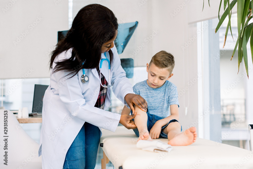 African woman doctor in a white coat carefully provides first aid. She examines the boy leg. The kid patiently observes the process, sitting on the couch. African woman doctor in a white coat carefully provides first aid. She examines the boy leg. The kid patiently observes the process, sitting on the couch.