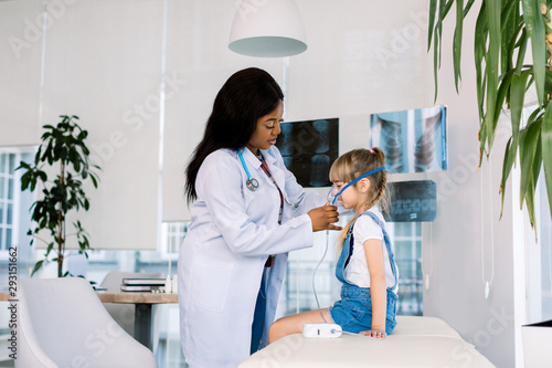 African woman doctor holds a mask vapor inhaler for little girl sitting on the couch. treatment of asthma. breathing through a steam nebulizer. concept of inhalation therapy apparatus photo