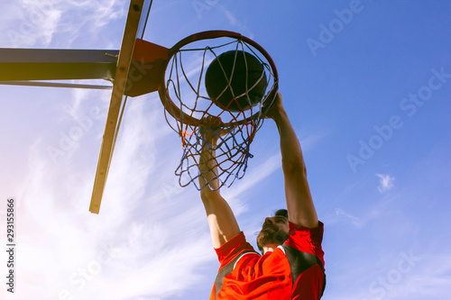 Bottom view of an athlete performing a dunk in a basketball hoop in a jump on a background of blue sky and bright sun. Sports lifestyle, desire for victory and success. Young guy basketball player.