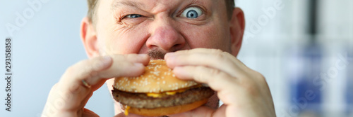 Funny bearded man with idiot facial expression eating big burger with enjoyment portrait