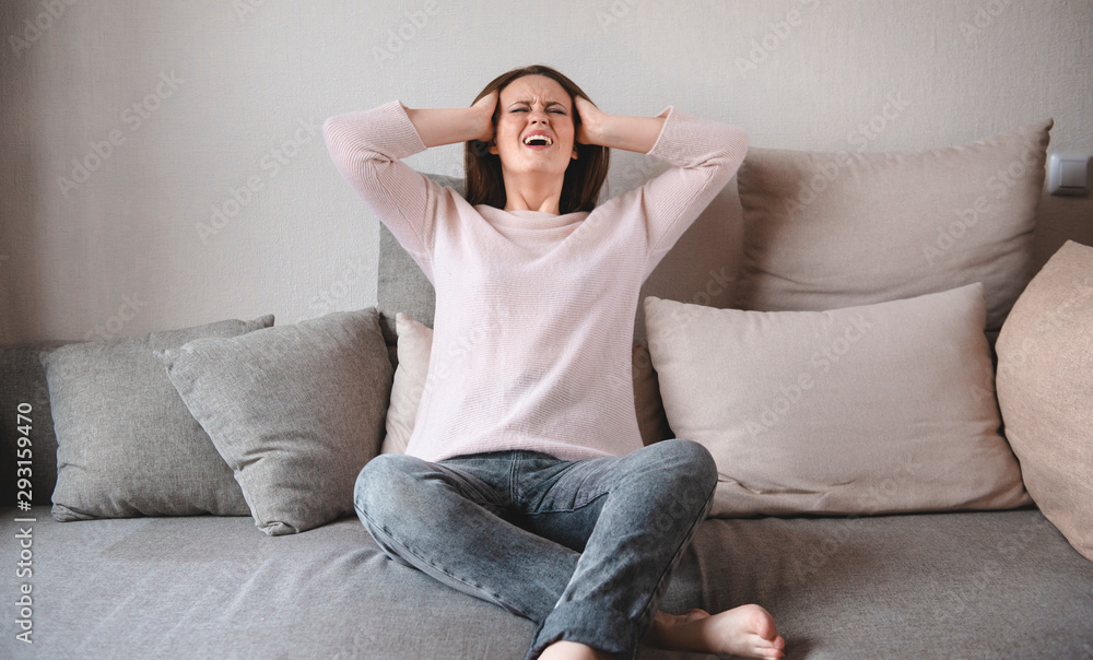 1 white young woman in pink sweater and grey jeans sitting on sofa holding her head in agony from headache, expression of agony on face 1 white young woman in pink sweater and grey jeans sitting on sofa holding her head in agony from headache, expression of agony on face