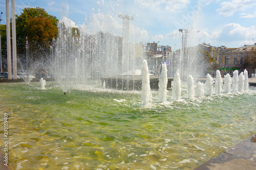 Waterworks fountain with water sprays and geysers on city park or street. Autumn day time freshness and relax concept. Clear aqua pool and yellow and red leaves trees