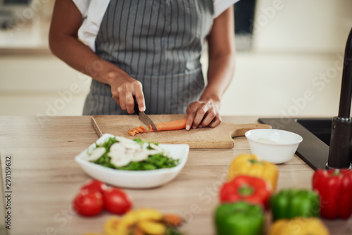 Close up of mixed race woman in apron cutting carrot for meal. On kitchen counter are vegetables.