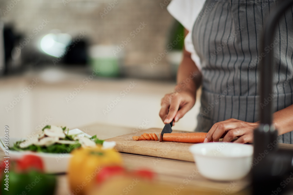 Close up of mixed race woman in apron cutting carrot for meal. On kitchen counter are vegetables.