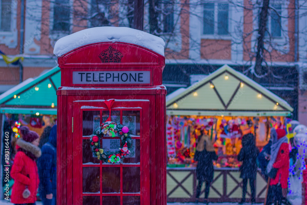 winter holidays and Christmas fair season celebration concept picture of red phone box foreground and shopping houses with unfocused people background in snowing weather time winter holidays and Christmas fair season celebration concept picture of red phone box foreground and shopping houses with unfocused people background in snowing weather time