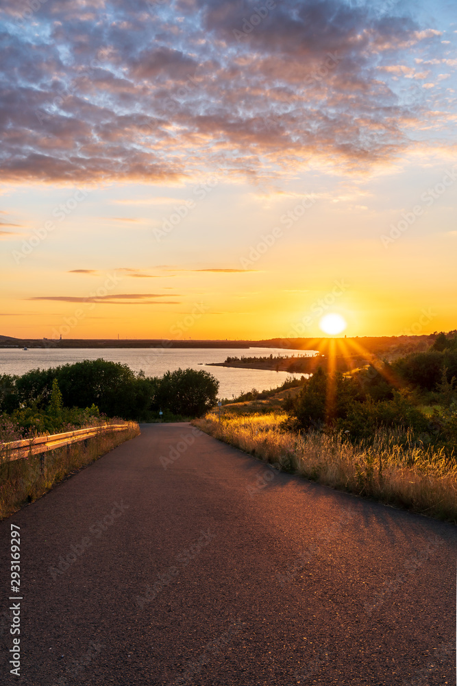 Fototapeta premium Asphalt road leading to a lake during a beautiful summer sunset 