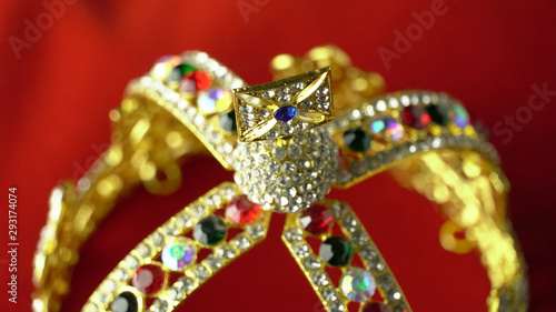 Gold diamond crown or decorative pageant accessory close up focus on the top. On the red royal color surface.
