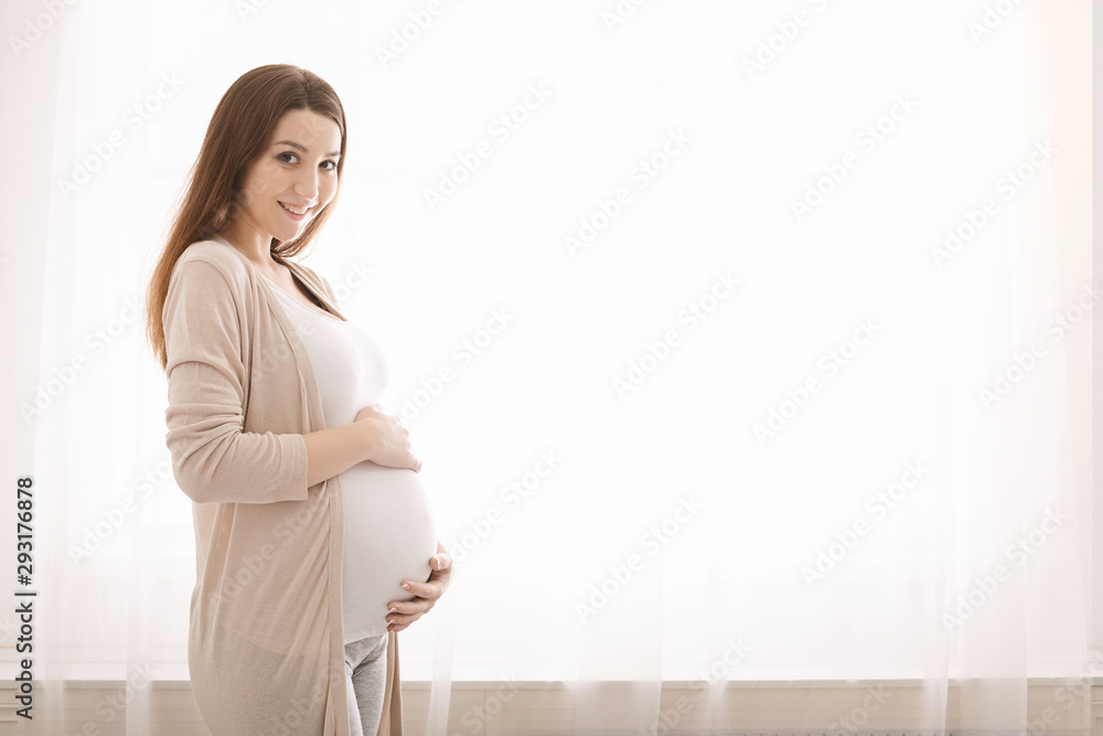 Young pregnant woman embracing her belly, standing near window - obrazy, fototapety, plakaty 