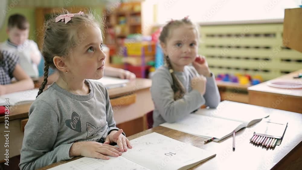 Twin girls in identical clothes, sitting at a desk in a preschool class. Children at school are sitting at the tables. Twin girls in identical clothes, sitting at a desk in a preschool class. Children at school are sitting at the tables.