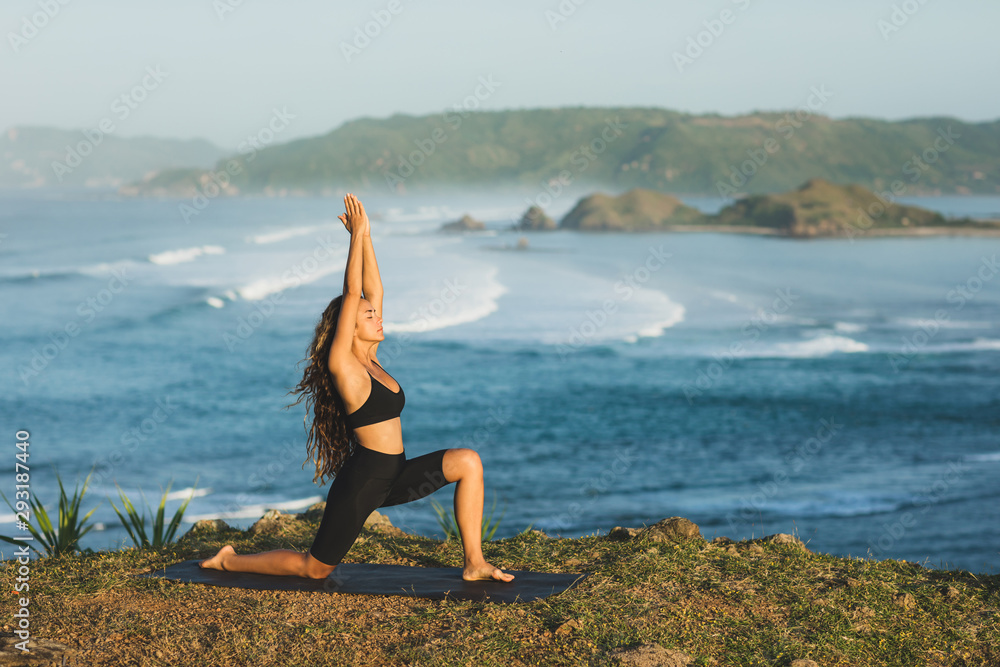 Woman practicing yoga outdoors with amazing ocean and mountain view in morning. Female health and fitness concept. Nature background.