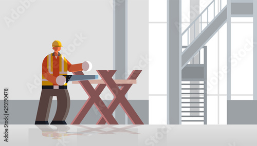 male builder carpenter using handsaw sawing log on sawbuck into lumber busy workman in uniform building concept construction site interior flat full length horizontal