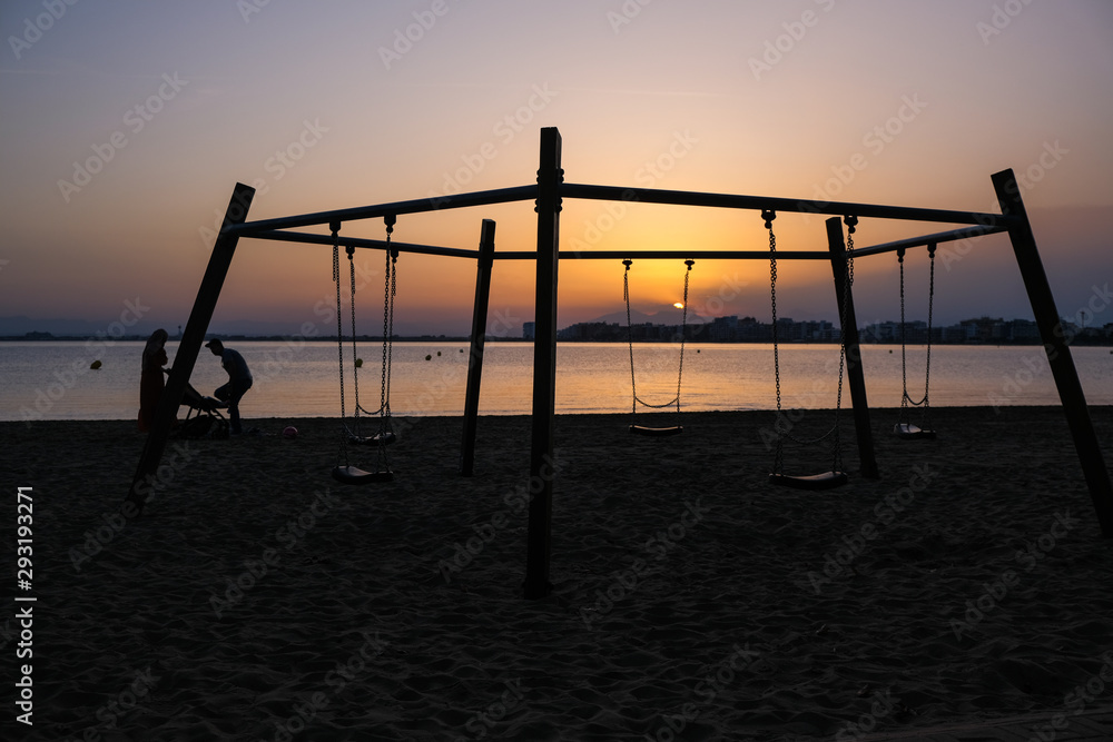 Children's swing on the beach on a background of sunset and sea. Silhouettes of a family of two parents and a child. Concept of family vacation. Summer sunset in Spain, Catalunya. Playground on beach. Children's swing on the beach on a background of sunset and sea. Silhouettes of a family of two parents and a child. Concept of family vacation. Summer sunset in Spain, Catalunya. Playground on beach.