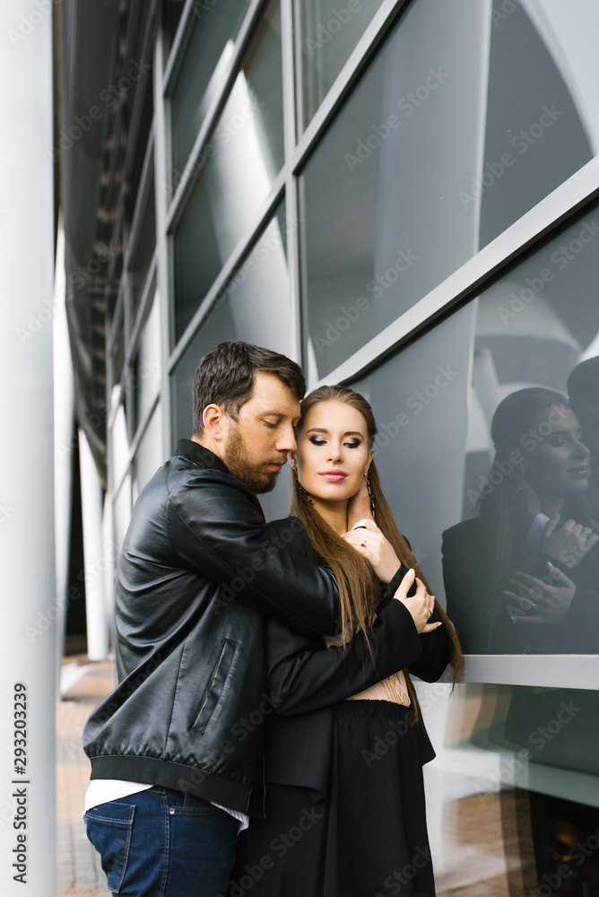 The guy hugs the girl neck, the girl closed her eyes in pleasure. Couple in black stylish clothes near the glass wall The guy hugs the girl neck, the girl closed her eyes in pleasure. Couple in black stylish clothes near the glass wall