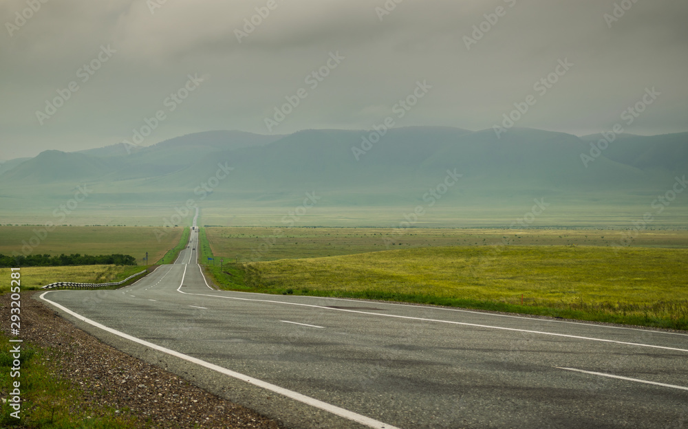 Highway road in Khakassia with green grass steppe and sayan mountains in fog and gray clouds in summer day, Siberia, Russia
