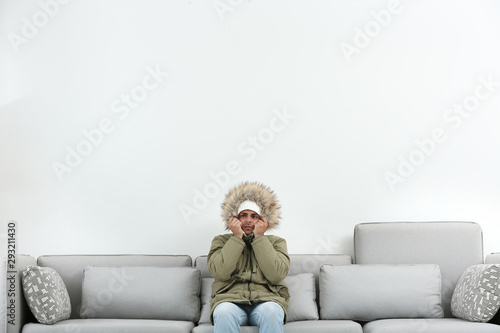 Tablou pe pânză Young man in warm clothes freezing on sofa against white background