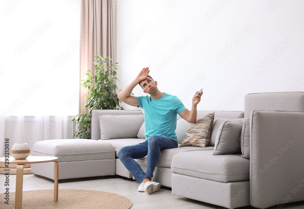 Young man with remote control suffering from heat under broken air conditioner at home Young man with remote control suffering from heat under broken air conditioner at home