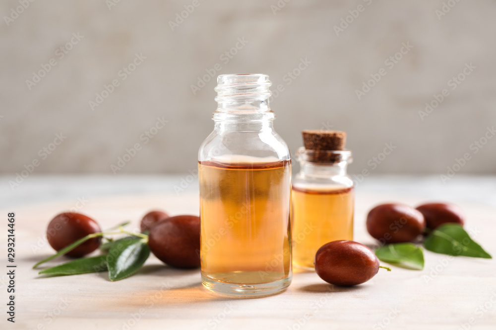 Glass bottles with jojoba oil and seeds on light table against grey background Glass bottles with jojoba oil and seeds on light table against grey background