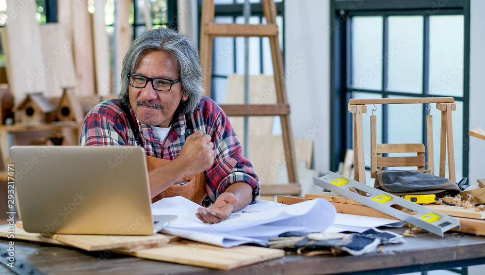 Old Asian craftsman act as feel happy with something appear on laptop screen and sit in his workplace. Concept of good work and income after retire and work at home with family. Old Asian craftsman act as feel happy with something appear on laptop screen and sit in his workplace. Concept of good work and income after retire and work at home with family.