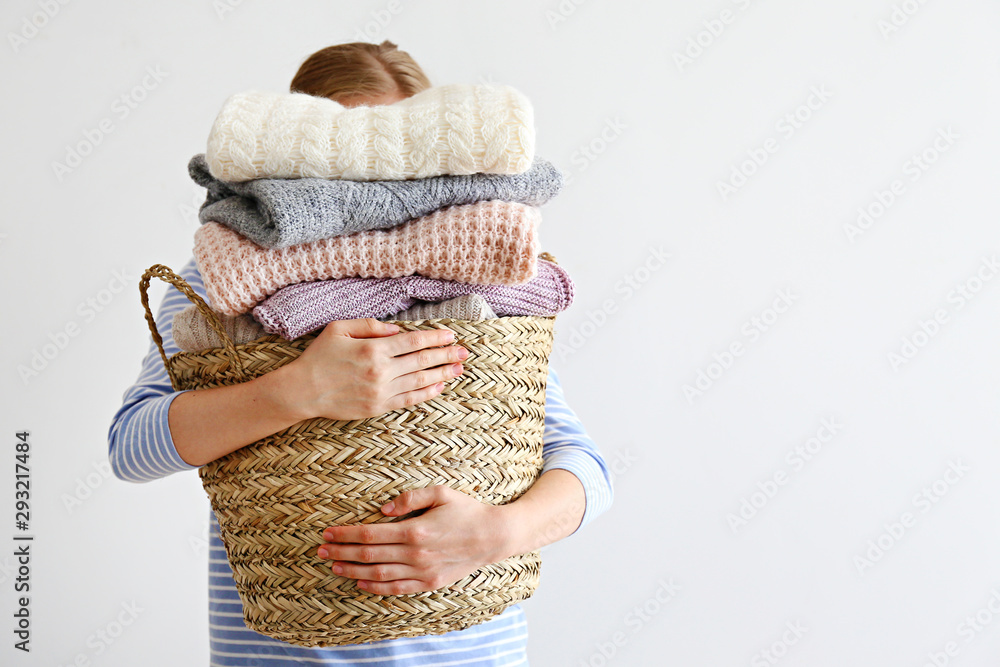 Slim young woman wearing oversized cotton shirt with blue stripes & holding big stack of folded knitted warm pastel color sweaters, easy chic style, different knitting patterns. Background, copy space
