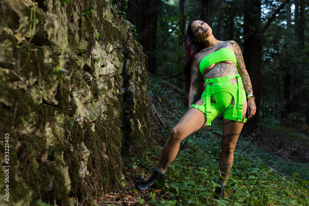 Asian tattooed woman in neon rave clothing poses on an abandoned rock wall in the forest Asian tattooed woman in neon rave clothing poses on an abandoned rock wall in the forest
