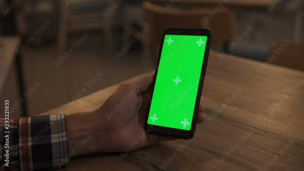 Close-up of a Man's Hand Holding Green Mock-up Screen Smartphone. Using app. Modern Mobile Phone. In the Background Cozy Living Room or Home Office. Close-up of a Man's Hand Holding Green Mock-up Screen Smartphone. Using app. Modern Mobile Phone. In the Background Cozy Living Room or Home Office.