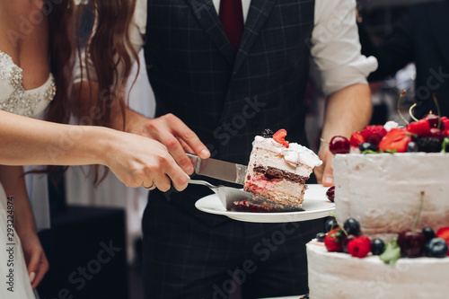 Side view of young bride and groom keeping knife and plate and cutting delicious wedding cake in restaurant. Piece of yummy sweet dessert. Concept of confectionery  celebration and love.