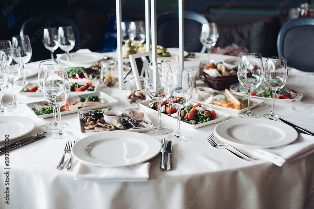 Fototapeta premium View over wedding table covered with white tablecloth with empty glasses, plates and utensils. Snacks are served on the table.