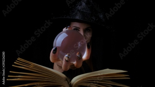 Wallpaper Mural Attractive mystic witch woman with bright red lips on a black background, looks into a magic ball while casting spells next to a spell book Torontodigital.ca