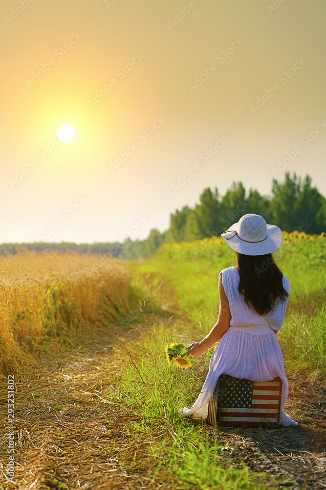 Rear view of girl with sunflowers, sitting on american flag suitcase, looking at sunset in golden wheat field Rear view of girl with sunflowers, sitting on american flag suitcase, looking at sunset in golden wheat field