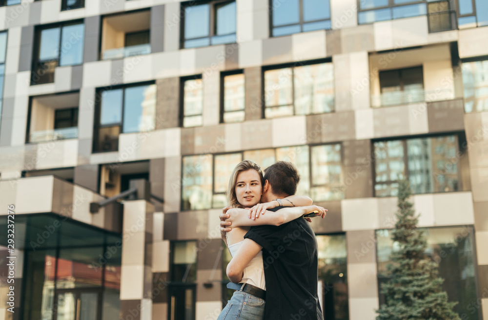 Beautiful young couple hugging on the street on the background of a modern building, the girl looks away. Guy hugged the girl on a walk on a warm summer day, she is happy. Love story