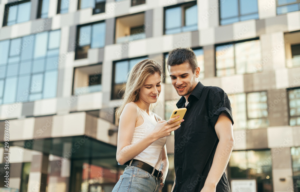 Beautiful young couple is standing on a background of modern architecture, looking at the screen of a smart phone and smiling. Happy girl and stylish young man using smartphone on the street.