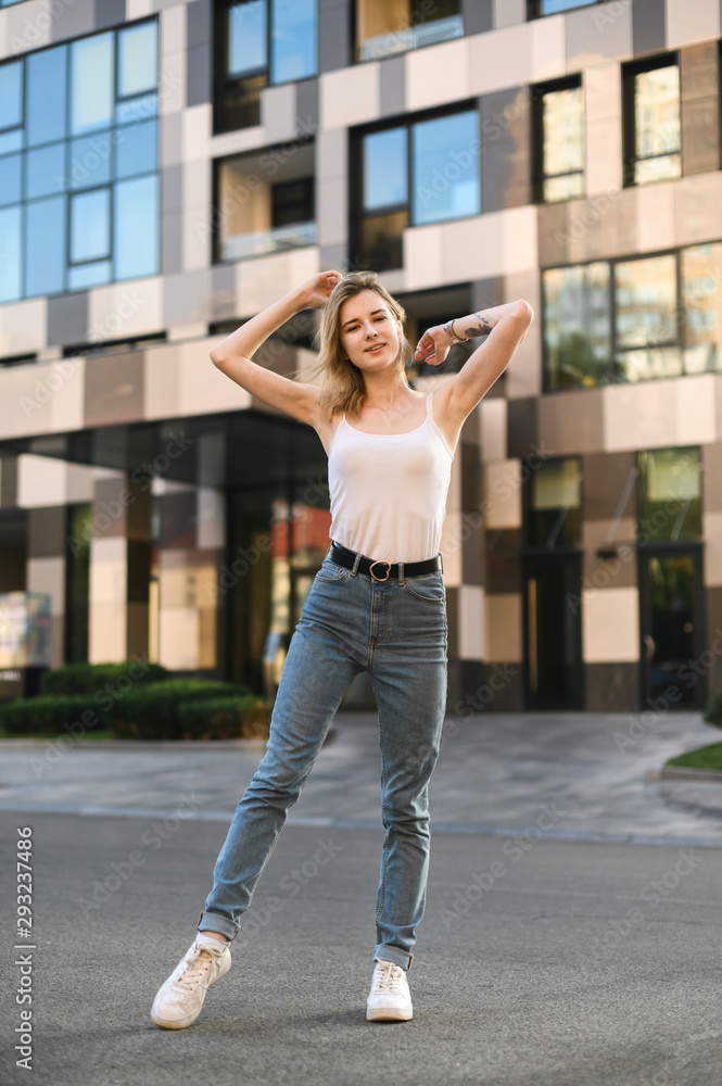 Full length portrait of attractive girl in casual clothes standing on the street and posing at camera, smiling. Girl with street style posing on the street.
