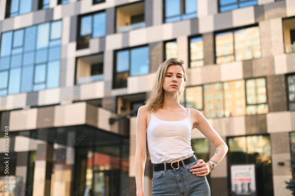 Street portrait of attractive girl in casual clothes standing on the street and posing, looking in the camera,smiling.Girl with street style posing on the street on a background of modern architecture