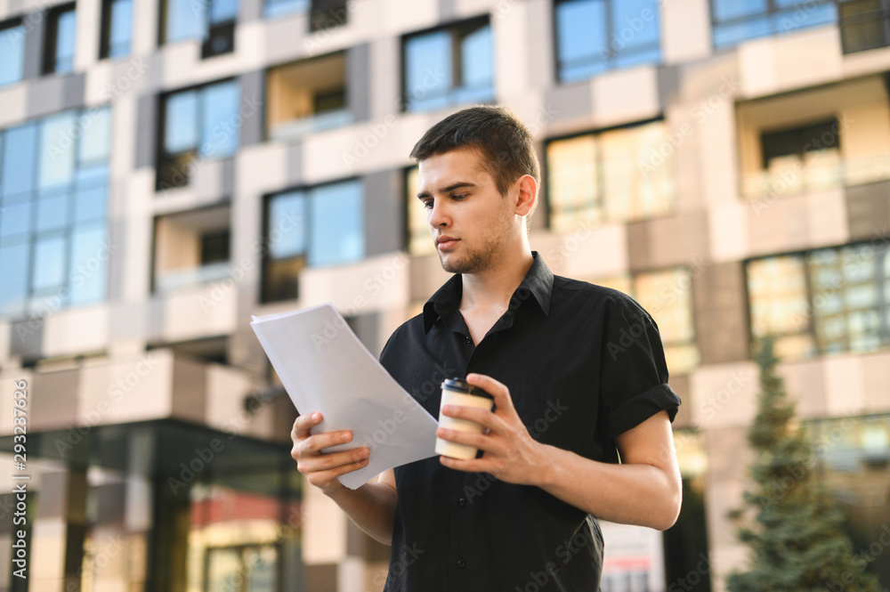 A street portrait of a young business man in a dark shirt stands on the street with documents and a cup of coffee in his hands, reading a serious face. Busy office worker studies papers on a walk.