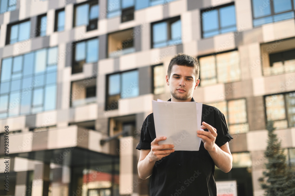 A business young man in a black shirt stands with papers in his hands and reads on the background of modern architecture. Serious guy with documents in his hands stands on the street, portrait.