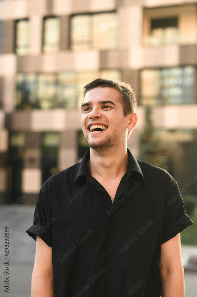 Smiling male model in stylish casual clothes posing at camera against the backdrop of a modern building looking upside down with a positive face. Handsome guy standing on urban background and smiling Smiling male model in stylish casual clothes posing at camera against the backdrop of a modern building looking upside down with a positive face. Handsome guy standing on urban background and smiling