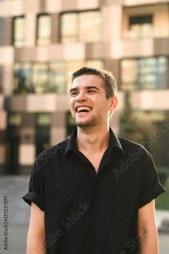 Smiling male model in stylish casual clothes posing at camera against the backdrop of a modern building looking upside down with a positive face. Handsome guy standing on urban background and smiling