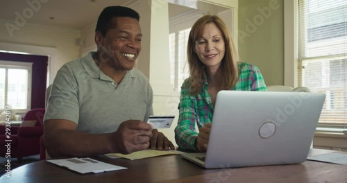 Happy older couple booking trip or making purchase online with credit card using laptop computer. African American and Caucasian husband and wife paying off outstanding debt. Slow motion 4k handheld