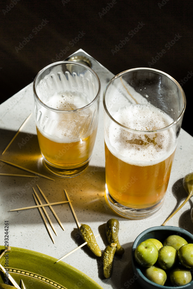 Beer and snacks. A selection of pickled gherkins, caper berries and green olives accompany two glasses of beer on a concrete background. Beer and snacks. A selection of pickled gherkins, caper berries and green olives accompany two glasses of beer on a concrete background.