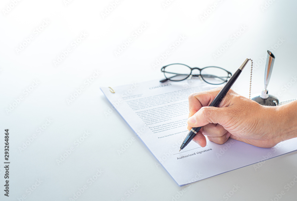 © Supagrit Ninkaesorn - White table with paper contract detail and empty space to sign authorized signature, props with glasses and ink pen, Hand holding object, copy space available for text