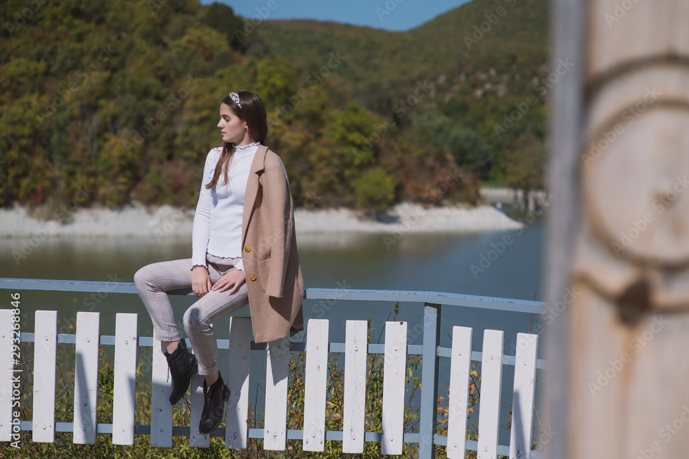 a beautiful, young brown-haired woman in a beige coat and a white turtleneck poses against a lake with cypresses in dark ankle boots