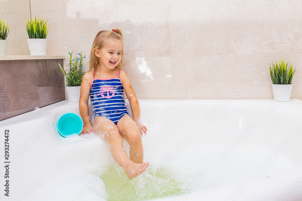 Little smiling blonde girl in blue swimsuit splashing in a big modern bathroom with foam. .Kids hygiene. Shampoo, hair treatment and soap for children. Little smiling blonde girl in blue swimsuit splashing in a big modern bathroom with foam. .Kids hygiene. Shampoo, hair treatment and soap for children.