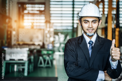 Engineer or business man wearing helmet standing and showing thumb up in front of heavy duty factory.